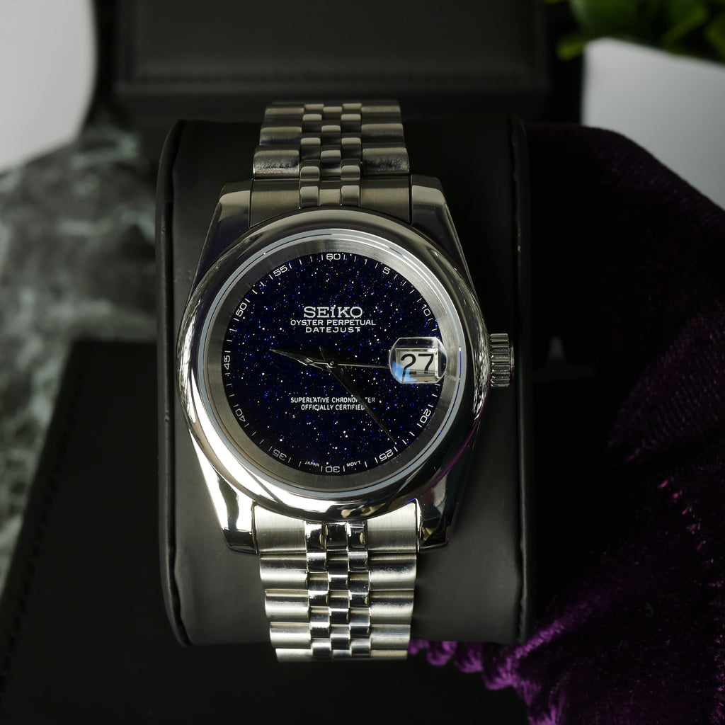 Silver watch with a dark face on a black stand with a purple cloth