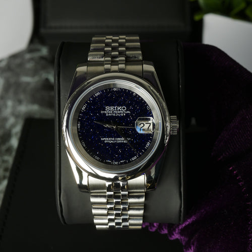 Silver watch with a dark face on a black stand with a purple cloth