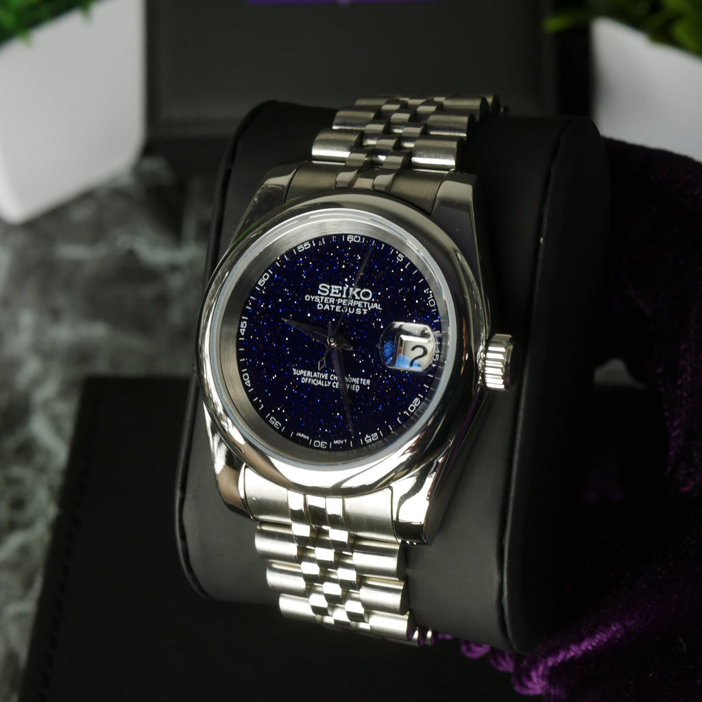 Silver Seiko watch with blue face on a black stand with blurred greenery in the background