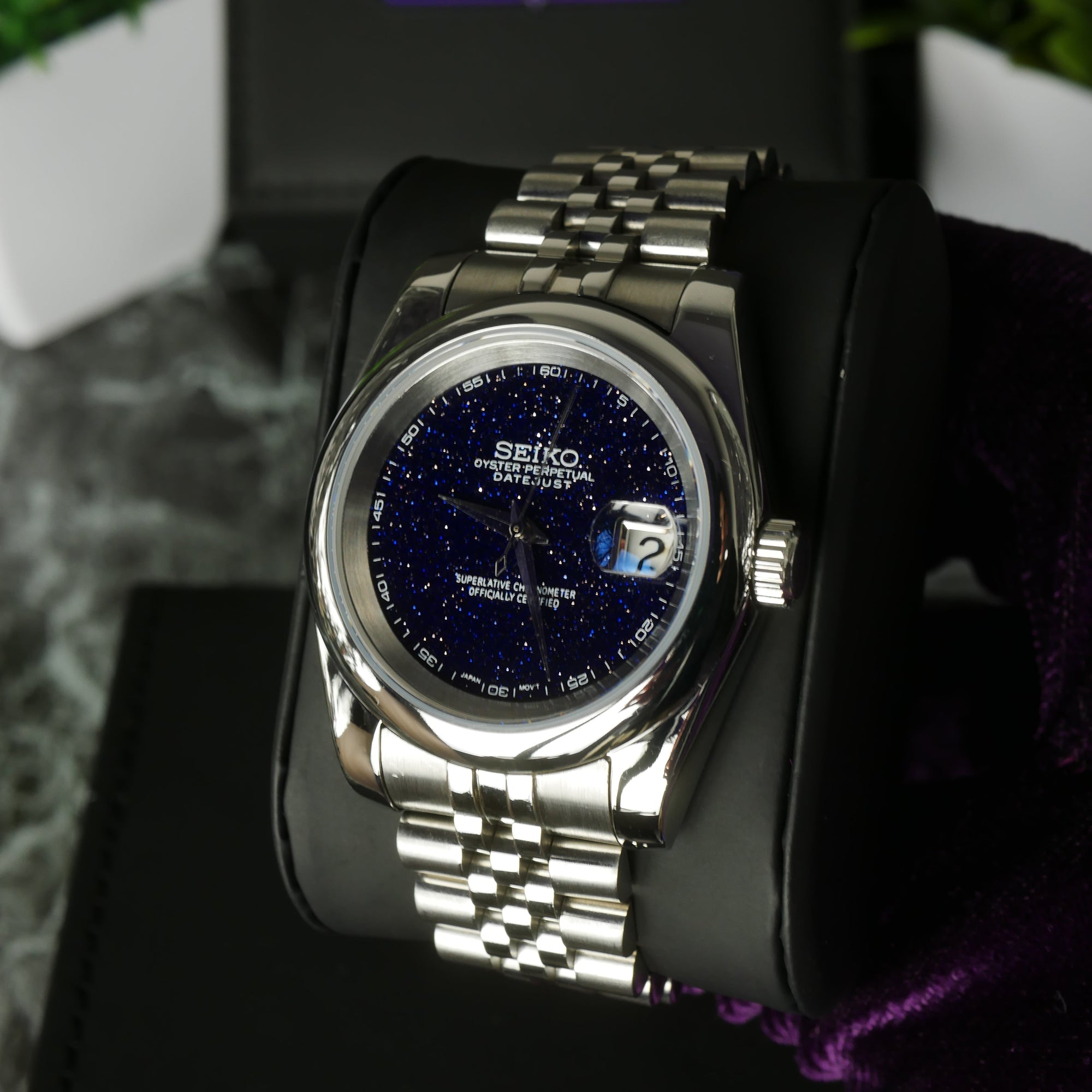 Silver Seiko watch with blue face on a black stand with blurred greenery in the background