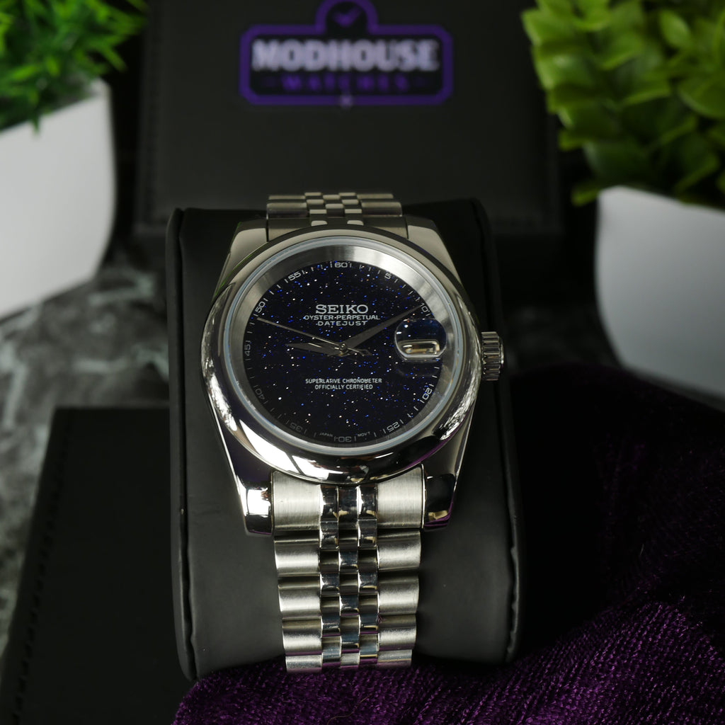 Silver Seiko watch on a black surface with a purple logo in the background